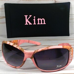 May include: Pink and brown camouflage sunglasses with rhinestones on the arms. The sunglasses have the name "Kim" written on the right lens.