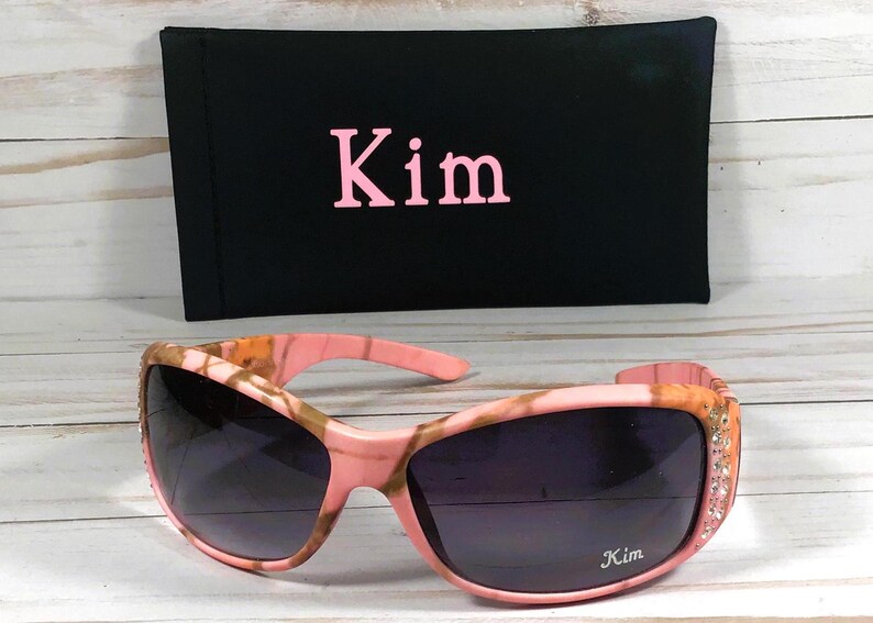 May include: Pink and brown camouflage sunglasses with rhinestones on the arms. The sunglasses have the name "Kim" written on the right lens.