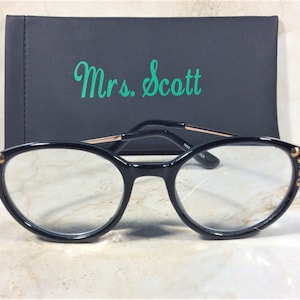 Pu&ograve; includere: Occhiali neri di forma ovale con dettagli dorati e borchie sulle montature. Gli occhiali sono davanti a un astuccio nero con il nome "Mrs. Scott" in verde.