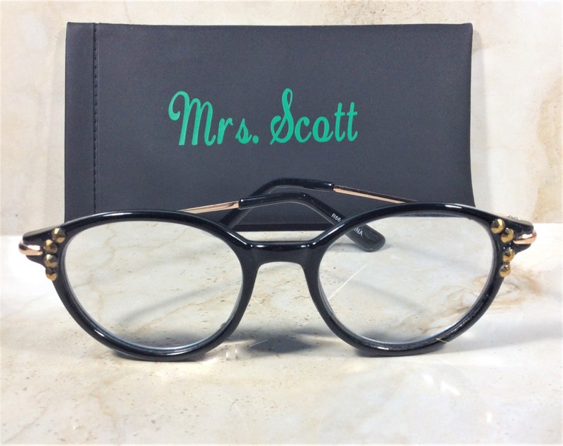 Pu&ograve; includere: Occhiali neri di forma ovale con dettagli dorati e borchie sulle montature. Gli occhiali sono davanti a un astuccio nero con il nome "Mrs. Scott" in verde.