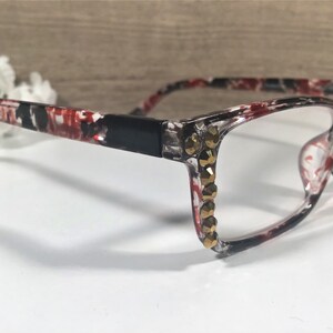 May include: Eyeglasses with a black, red, and white patterned frame. The frames have clear lenses and are embellished with gold-colored rhinestones along the outer edges, adding a touch of glamour to the eyewear.