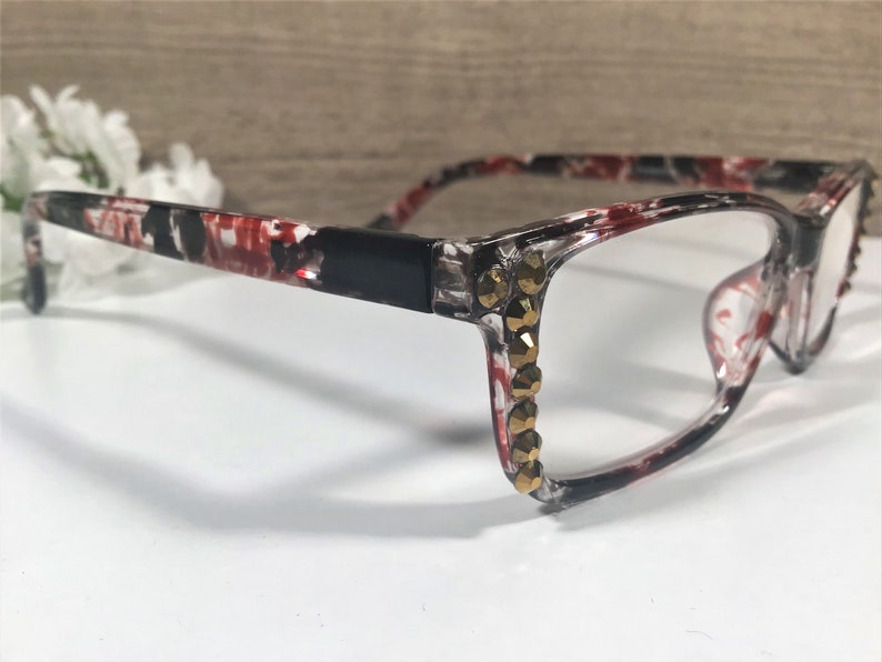 May include: Eyeglasses with a black, red, and white patterned frame. The frames have clear lenses and are embellished with gold-colored rhinestones along the outer edges, adding a touch of glamour to the eyewear.