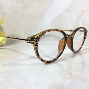 May include: Round eyeglasses with a tortoiseshell pattern frame and gold-tone metal arms. The frame is embellished with small, gold-colored studs. The lenses are clear. A yellow flower is in the background.