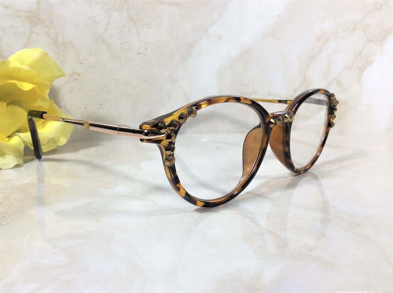 May include: Round eyeglasses with a tortoiseshell pattern frame and gold-tone metal arms. The frame is embellished with small, gold-colored studs. The lenses are clear. A yellow flower is in the background.