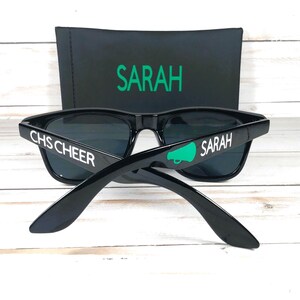 Puede incluir: Gafas de sol negras con lentes oscuras y texto blanco que dice "CHS CHEER" y "SARAH". Un gráfico de megáfono verde está en una patilla. Una funda negra con "SARAH" en verde está detrás de las gafas.