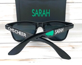 Może przedstawiać: Czarne okulary przeciwsłoneczne z ciemnymi soczewkami i białym napisem "CHS CHEER" i "SARAH". Zielona grafika megafonu znajduje się na jednym zauszniku. Czarne etui z napisem "SARAH" w kolorze zielonym znajduje się za okularami.