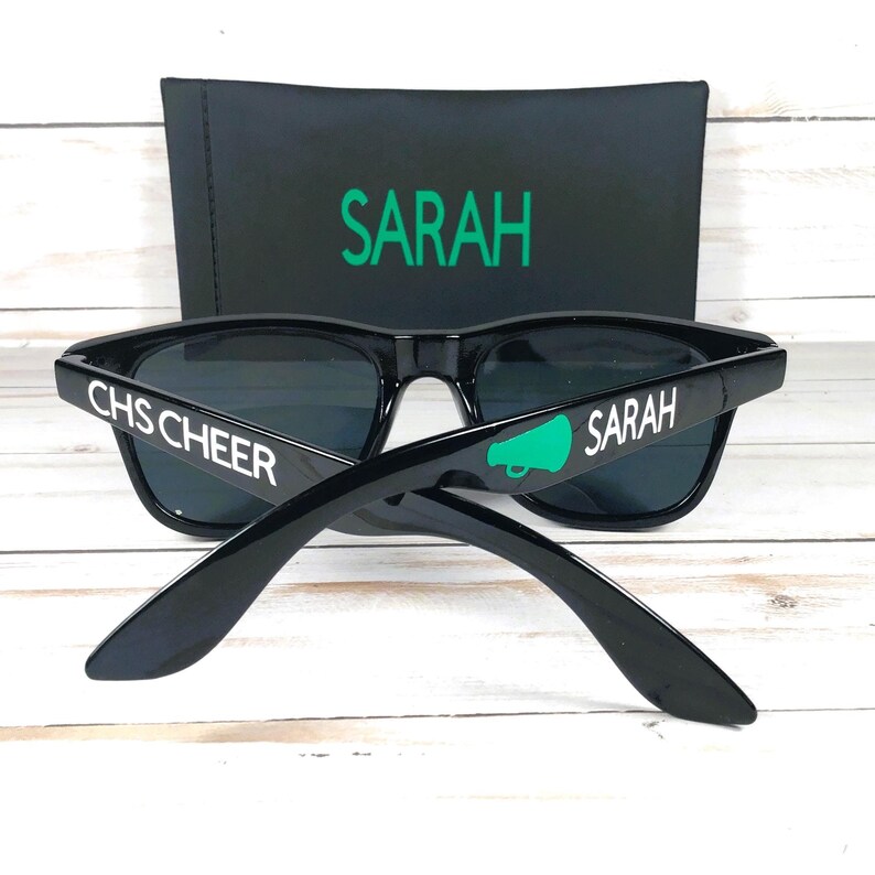 May include: Black sunglasses with dark lenses and white text that reads "CHS CHEER" and "SARAH". A green megaphone graphic is on one arm. A black case with "SARAH" in green is behind the sunglasses.