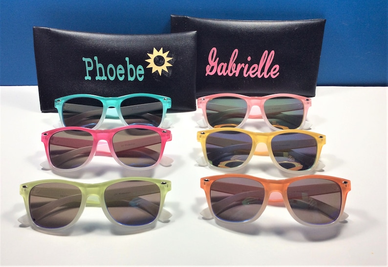May include: Six pairs of colorful sunglasses with gradient lenses. The sunglasses are arranged in two rows of three. The sunglasses are in the colors of turquoise, pink, yellow, and orange. The sunglasses are on a white surface. The sunglasses are in a black case with the names "Phoebe" and "Gabrielle" written on them.