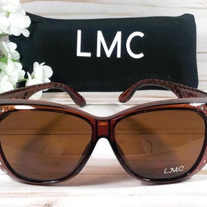 May include: Brown sunglasses with a dark brown frame and lenses. The frame has a decorative pattern on the temples and is embellished with small rhinestones. The sunglasses are paired with a black case with the letters "LMC" in white.