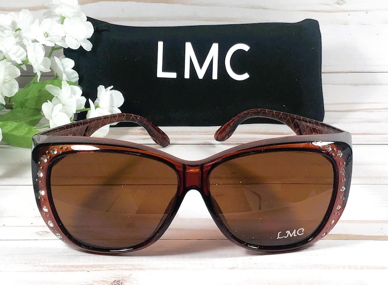 May include: Brown sunglasses with a dark brown frame and lenses. The frame has a decorative pattern on the temples and is embellished with small rhinestones. The sunglasses are paired with a black case with the letters "LMC" in white.