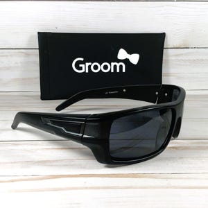Pu&ograve; includere: Occhiali da sole neri con montatura rettangolare e lenti scure. Gli occhiali da sole sono accanto a un astuccio nero con la scritta "Groom" e un logo a farfallino bianco. "UV Protection" &egrave; stampato sulla montatura.