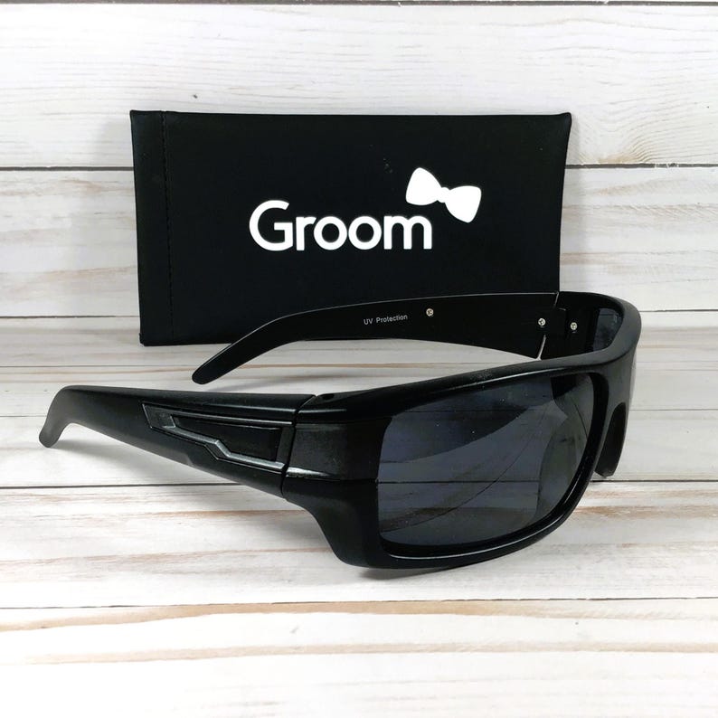 Pu&ograve; includere: Occhiali da sole neri con montatura rettangolare e lenti scure. Gli occhiali da sole sono accanto a un astuccio nero con la scritta "Groom" e un logo a farfallino bianco. "UV Protection" &egrave; stampato sulla montatura.