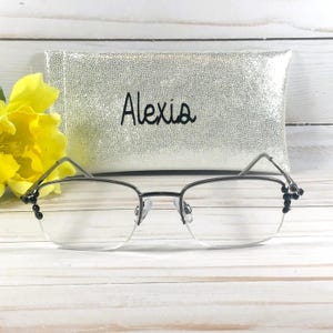 May include: A pair of silver-framed eyeglasses with clear lenses, resting in front of a silver sequined case. The case is embroidered with the name "Alexis" in black script. A yellow flower is visible on the left.