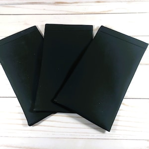 Puede incluir: Tres bolsas de tela negras, posiblemente para gafas u otros art&iacute;culos peque&ntilde;os, est&aacute;n apiladas sobre una superficie de madera blanca.