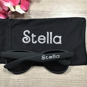 May include: Black sunglasses with the name "Stella" printed on the frame and a black carrying case with the name "Stella" printed on it.