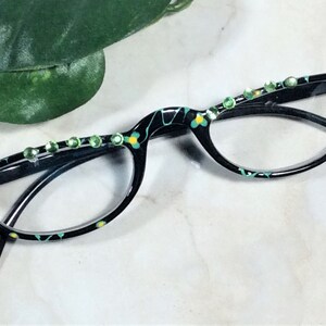 May include: Black eyeglasses with a floral pattern in teal and yellow. The frames are embellished with green rhinestones. The glasses have a cat-eye shape.