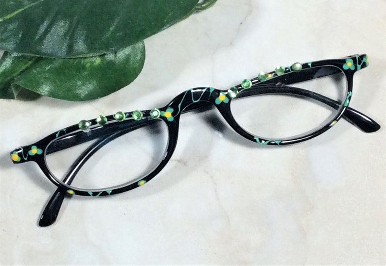 May include: Black eyeglasses with a floral pattern in teal and yellow. The frames are embellished with green rhinestones. The glasses have a cat-eye shape.