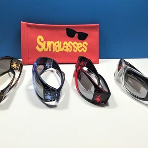 May include: Four pairs of sunglasses with spider web and spider designs. The sunglasses are in four different colours: orange, blue, red, and clear. The sunglasses are on a white surface with a red sunglasses case in the background. The case has the word "Sunglasses" printed on it in yellow letters.