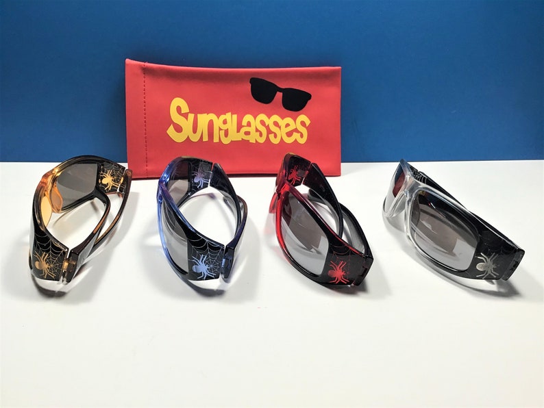 May include: Four pairs of sunglasses with spider web and spider designs. The sunglasses are in four different colours: orange, blue, red, and clear. The sunglasses are on a white surface with a red sunglasses case in the background. The case has the word "Sunglasses" printed on it in yellow letters.