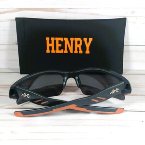 K&ouml;nnte beinhalten: Schwarze Sonnenbrille mit orangefarbenen Akzenten und einem schwarzen Etui mit dem Namen "HENRY" darauf.