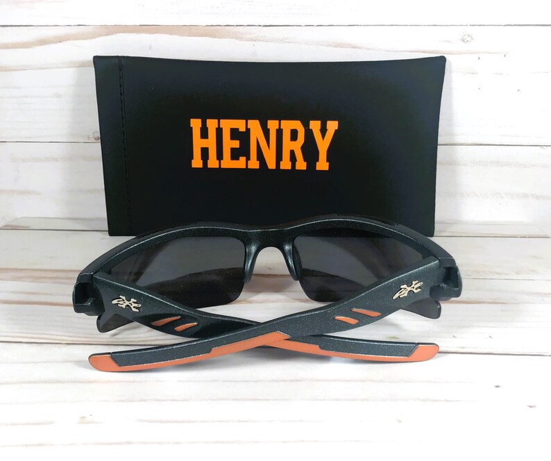K&ouml;nnte beinhalten: Schwarze Sonnenbrille mit orangefarbenen Akzenten und einem schwarzen Etui mit dem Namen "HENRY" darauf.