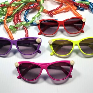 May include: Four pairs of colourful cat-eye sunglasses with white pearl accents. The sunglasses are in red, purple, yellow, and pink.