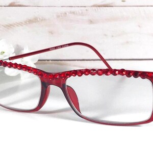 May include: Red rectangular eyeglasses with a decorative beaded top frame. The glasses have clear lenses and a matching red frame. The image shows the glasses on a white surface with white flowers in the background.