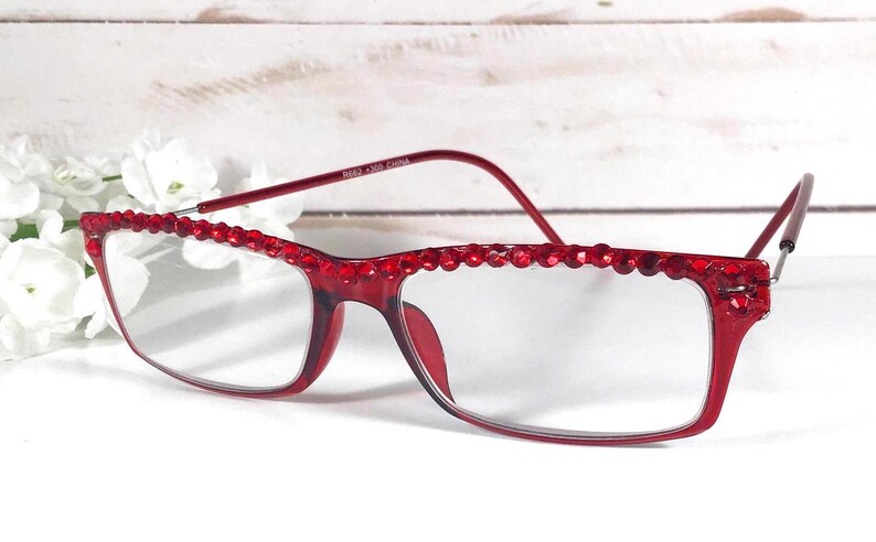 May include: Red rectangular eyeglasses with a decorative beaded top frame. The glasses have clear lenses and a matching red frame. The image shows the glasses on a white surface with white flowers in the background.