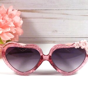 May include: Pink heart-shaped sunglasses with glitter and a pink flower detail on the side.