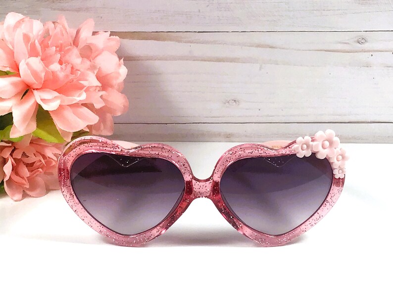 May include: Pink heart-shaped sunglasses with glitter and a pink flower detail on the side.