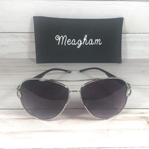 May include: Silver-framed aviator sunglasses with dark lenses. The sunglasses are paired with a black case, which has the name "Meagham" in white script. The sunglasses are on a light wood-grain surface.