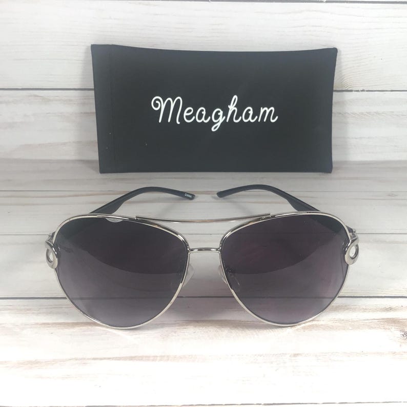 May include: Silver-framed aviator sunglasses with dark lenses. The sunglasses are paired with a black case, which has the name "Meagham" in white script. The sunglasses are on a light wood-grain surface.