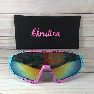 May include: Oversized sunglasses featuring a pink and light blue frame with a black paint splatter pattern and reflective rainbow lenses. Behind the sunglasses is a black rectangular case with the name Christina in pink script.