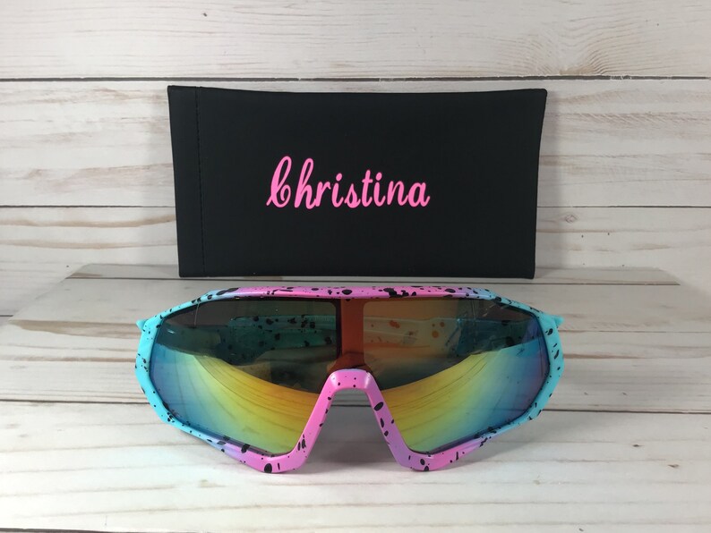 May include: Oversized sunglasses featuring a pink and light blue frame with a black paint splatter pattern and reflective rainbow lenses. Behind the sunglasses is a black rectangular case with the name Christina in pink script.