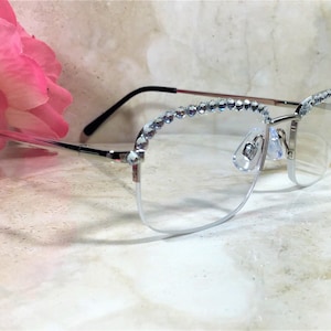 May include: Silver-framed eyeglasses with clear lenses. The top of the frames are embellished with a row of clear, sparkling rhinestones. The glasses have a half-rim design and are set against a marble background.
