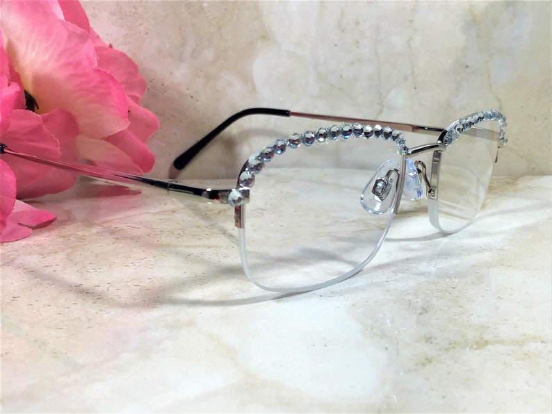 May include: Silver-framed eyeglasses with clear lenses. The top of the frames are embellished with a row of clear, sparkling rhinestones. The glasses have a half-rim design and are set against a marble background.