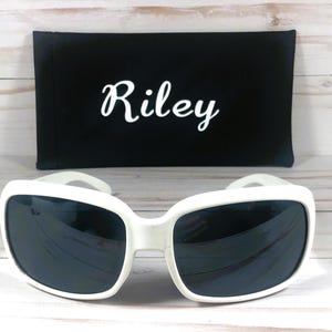 May include: White-framed sunglasses with dark lenses and a black case. The case has the name "Riley" in white script. The sunglasses have a square shape and a thick frame. The case is rectangular.
