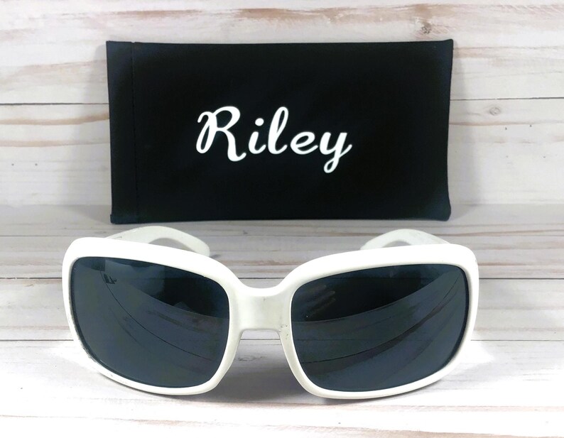 May include: White-framed sunglasses with dark lenses and a black case. The case has the name "Riley" in white script. The sunglasses have a square shape and a thick frame. The case is rectangular.