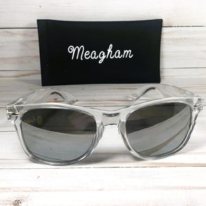 Op de afbeelding: Transparante zonnebril met spiegelglazen voor een zwarte etui. De naam "Meagham" is in witte letters op de etui geborduurd. De zonnebril heeft een klassiek, retro design.
