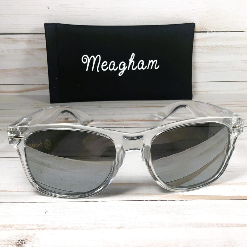 Op de afbeelding: Transparante zonnebril met spiegelglazen voor een zwarte etui. De naam "Meagham" is in witte letters op de etui geborduurd. De zonnebril heeft een klassiek, retro design.