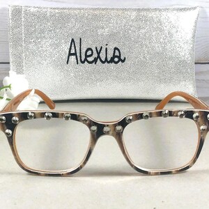 Puede incluir: Gafas con montura de estampado de leopardo y lentes transparentes, adornadas con tachuelas plateadas en la parte superior. Las gafas se combinan con un estuche plateado con el nombre "Alexia" en escritura negra.
