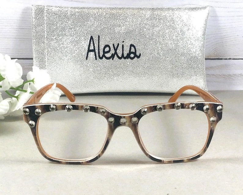 Puede incluir: Gafas con montura de estampado de leopardo y lentes transparentes, adornadas con tachuelas plateadas en la parte superior. Las gafas se combinan con un estuche plateado con el nombre "Alexia" en escritura negra.