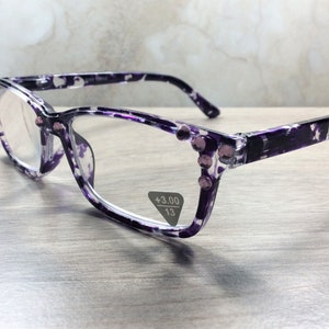 K&ouml;nnte beinhalten: Lesebrille mit lila-wei&szlig;em Blumenmuster. Die Fassungen haben eine Cat-Eye-Form und sind mit kleinen, rosa Strasssteinen verziert. Die Gl&auml;ser haben eine St&auml;rke von +3,00 und einen Steg von 13 mm.