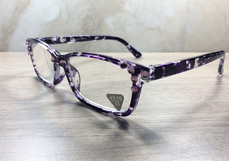 K&ouml;nnte beinhalten: Lesebrille mit lila-wei&szlig;em Blumenmuster. Die Fassungen haben eine Cat-Eye-Form und sind mit kleinen, rosa Strasssteinen verziert. Die Gl&auml;ser haben eine St&auml;rke von +3,00 und einen Steg von 13 mm.