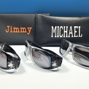 May include: Three pairs of black sunglasses with spider web designs and grey lenses. The sunglasses are in front of two black cases with the names "Jimmy" and "MICHAEL" printed on them.