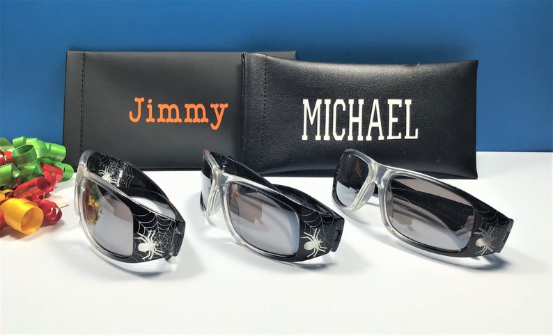 May include: Three pairs of black sunglasses with spider web designs and grey lenses. The sunglasses are in front of two black cases with the names "Jimmy" and "MICHAEL" printed on them.