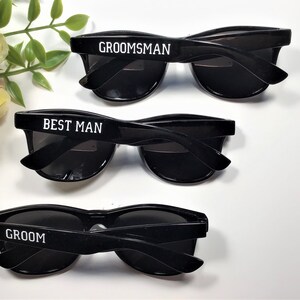 May include: Three pairs of black sunglasses with white text on the frames. The sunglasses say "Groom", "Best Man", and "Groomsman".