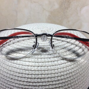Puede incluir: Gafas con montura plateada, detalles en rojo y adornos decorativos en las patillas. Las lentes son de media montura, y la montura incluye el texto "R375 +2.75" y "UV Protection". Las gafas est&aacute;n sobre una superficie blanca texturizada.