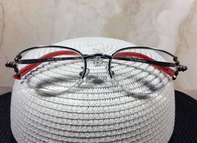 Puede incluir: Gafas con montura plateada, detalles en rojo y adornos decorativos en las patillas. Las lentes son de media montura, y la montura incluye el texto "R375 +2.75" y "UV Protection". Las gafas est&aacute;n sobre una superficie blanca texturizada.