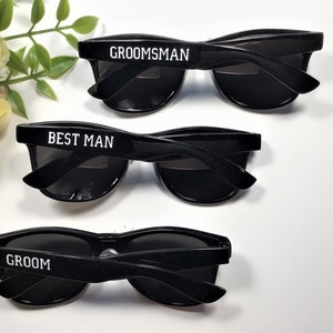 May include: Three pairs of black sunglasses with white text on the frames. The sunglasses say "Groom", "Best Man", and "Groomsman".